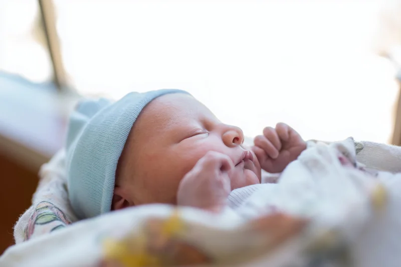 Newborn baby sleeping peacefully with eyes closed, wearing a light blue knitted hat, wrapped in a soft blanket near a window