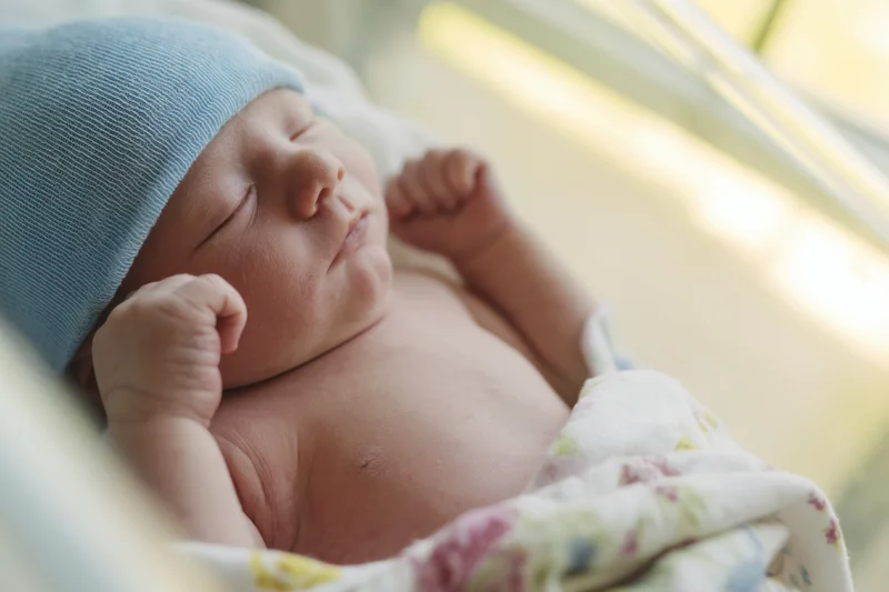 Newborn baby sleeping peacefully in a hospital crib wearing a light blue knit hat and wrapped in a floral blanket