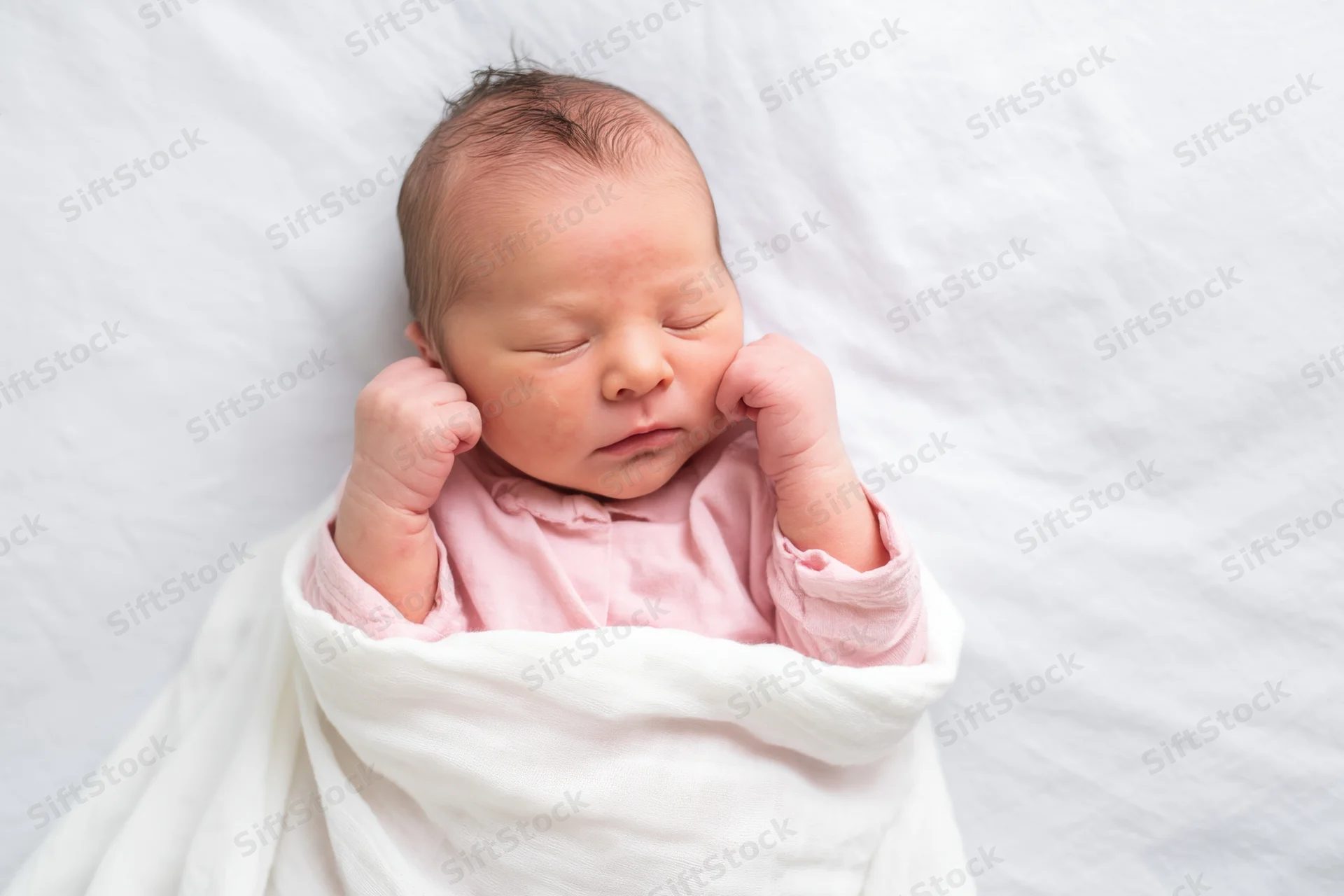 Newborn baby sleeping swaddled in white blanket with pink clothing, overhead view
