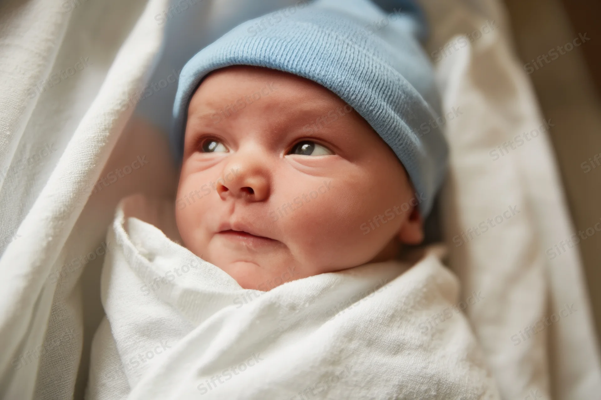 Newborn baby swaddled in white blanket wearing blue hat, close-up portrait