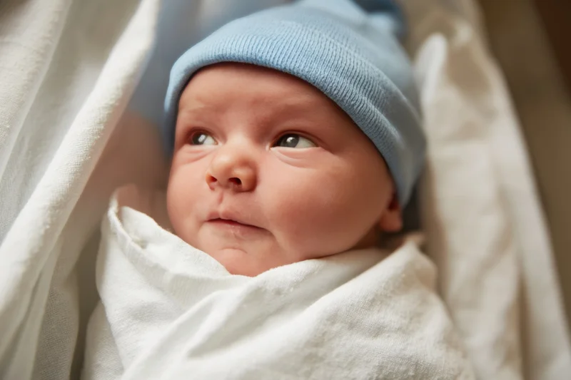 Newborn baby swaddled in white blanket wearing blue hat, close-up portrait