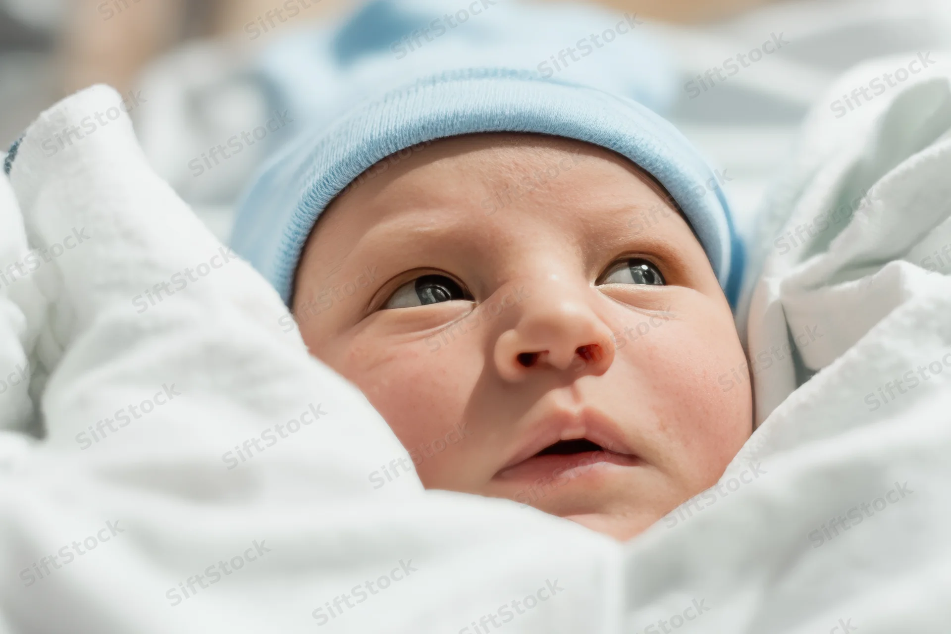 Newborn baby swaddled in white blankets wearing a blue knit hat, close-up of face looking upward
