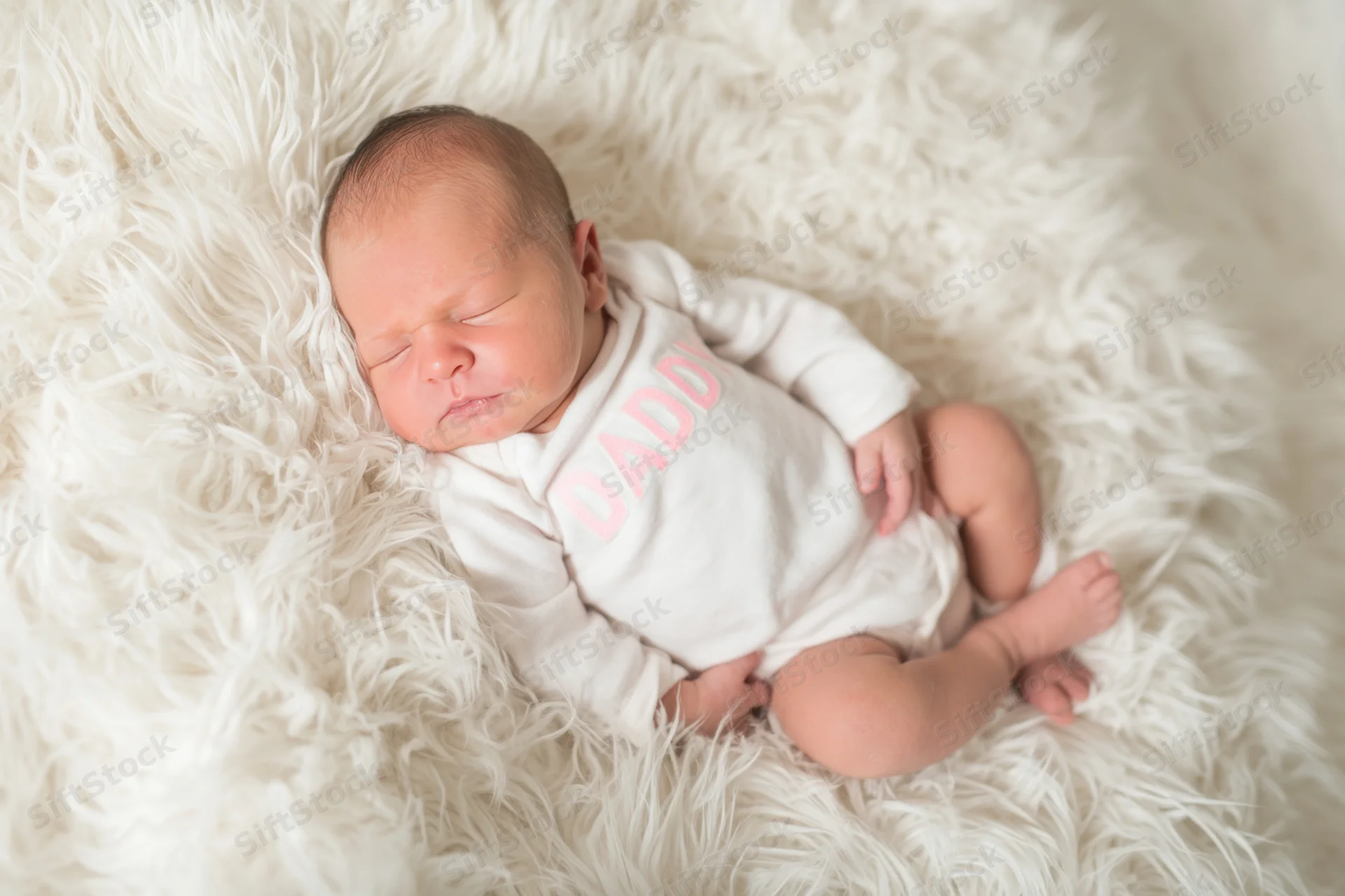 Newborn baby sleeping peacefully on a fluffy white fur blanket wearing a white onesie with pink "DADDY" text