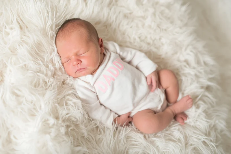 Newborn baby sleeping peacefully on a fluffy white fur blanket wearing a white onesie with pink "DADDY" text