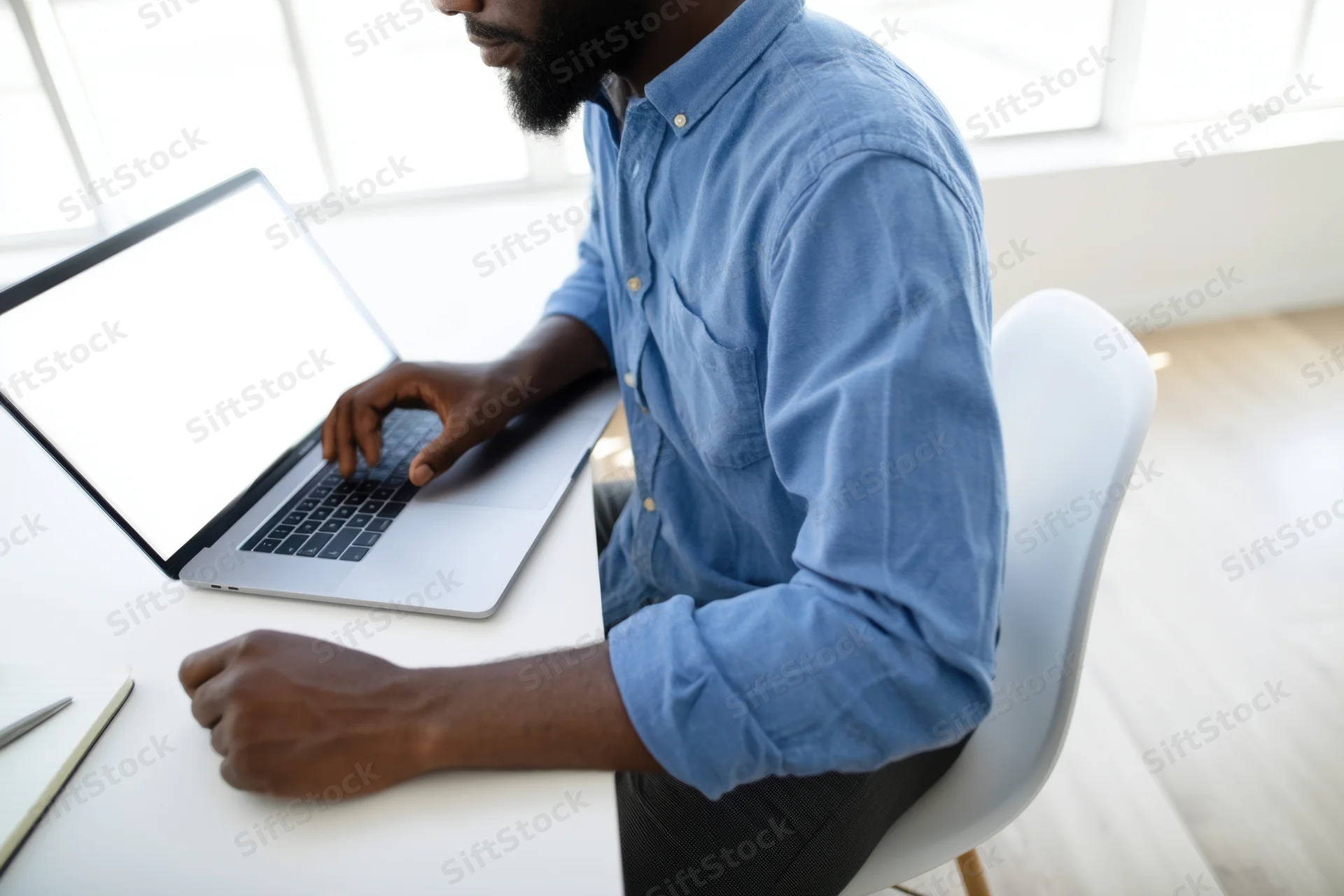 Man in blue shirt typing on laptop at white desk in bright office