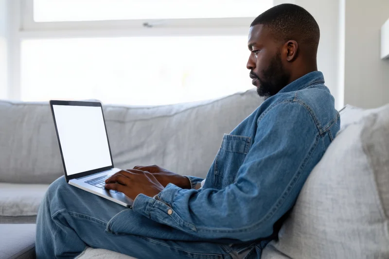 Man typing on laptop while sitting on light gray sofa in bright living room