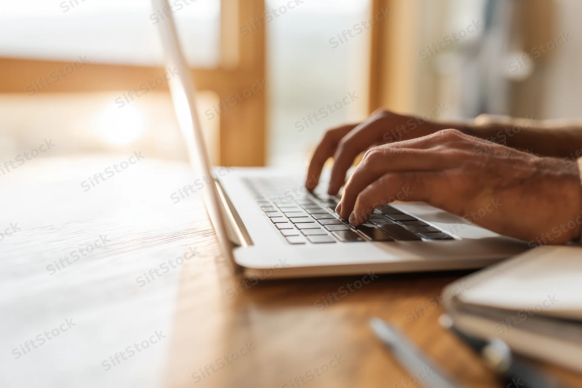 Hands typing on a sleek laptop at a wooden table in a sunlit room