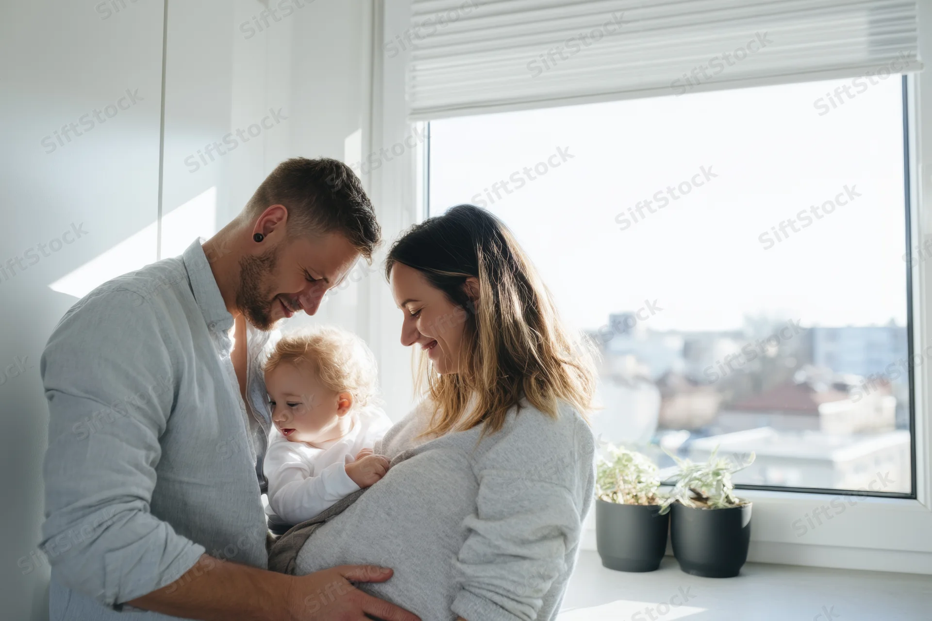 Pregnant woman and her partner holding baby together in bright modern kitchen by window