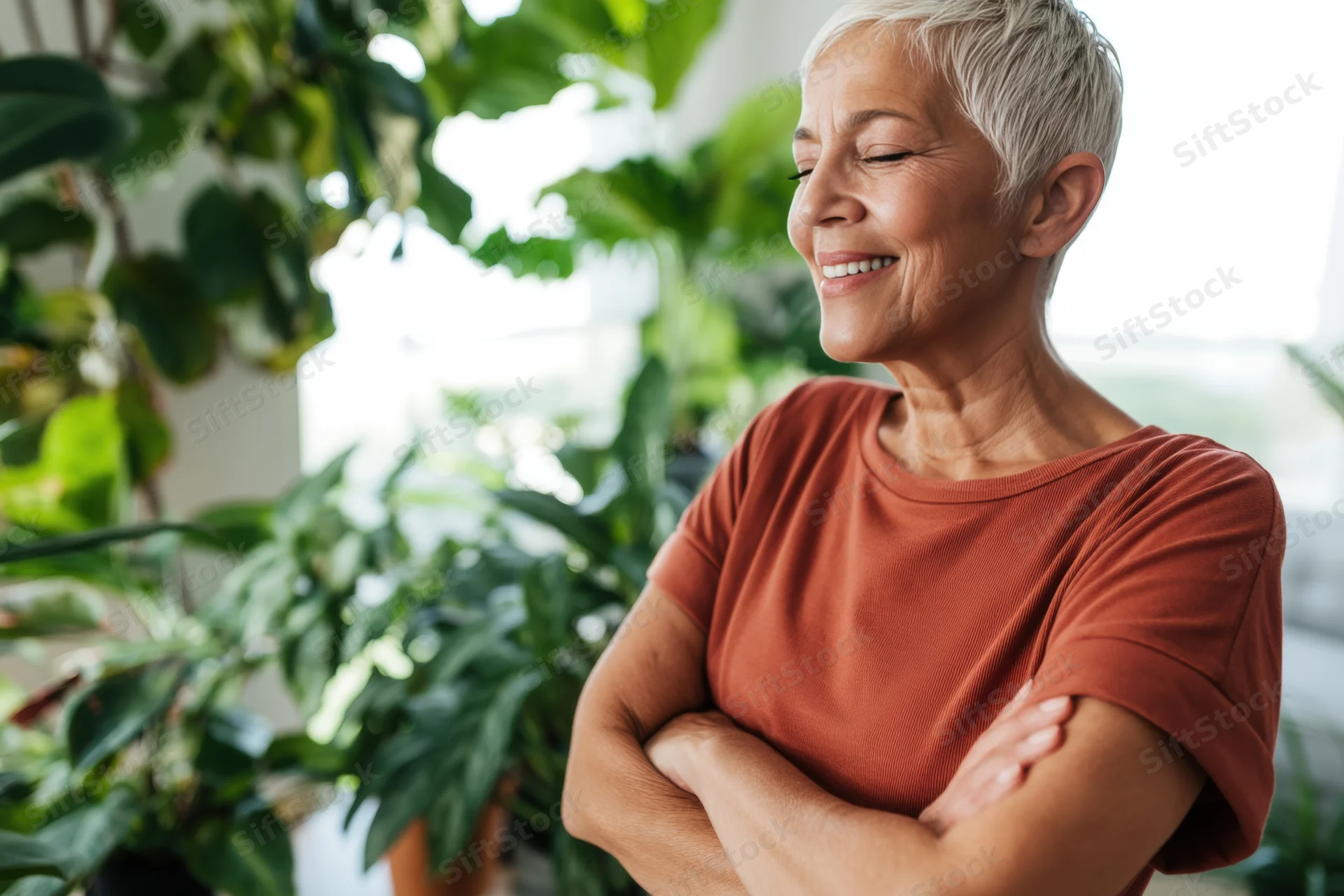 Senior woman smiling with arms crossed in a sunlit room filled with green houseplants