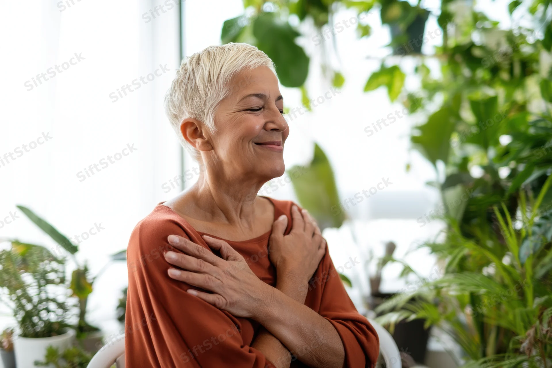Senior woman smiling with hands on chest surrounded by houseplants indoors
