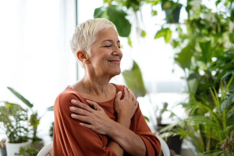 Senior woman smiling with hands on chest surrounded by houseplants indoors