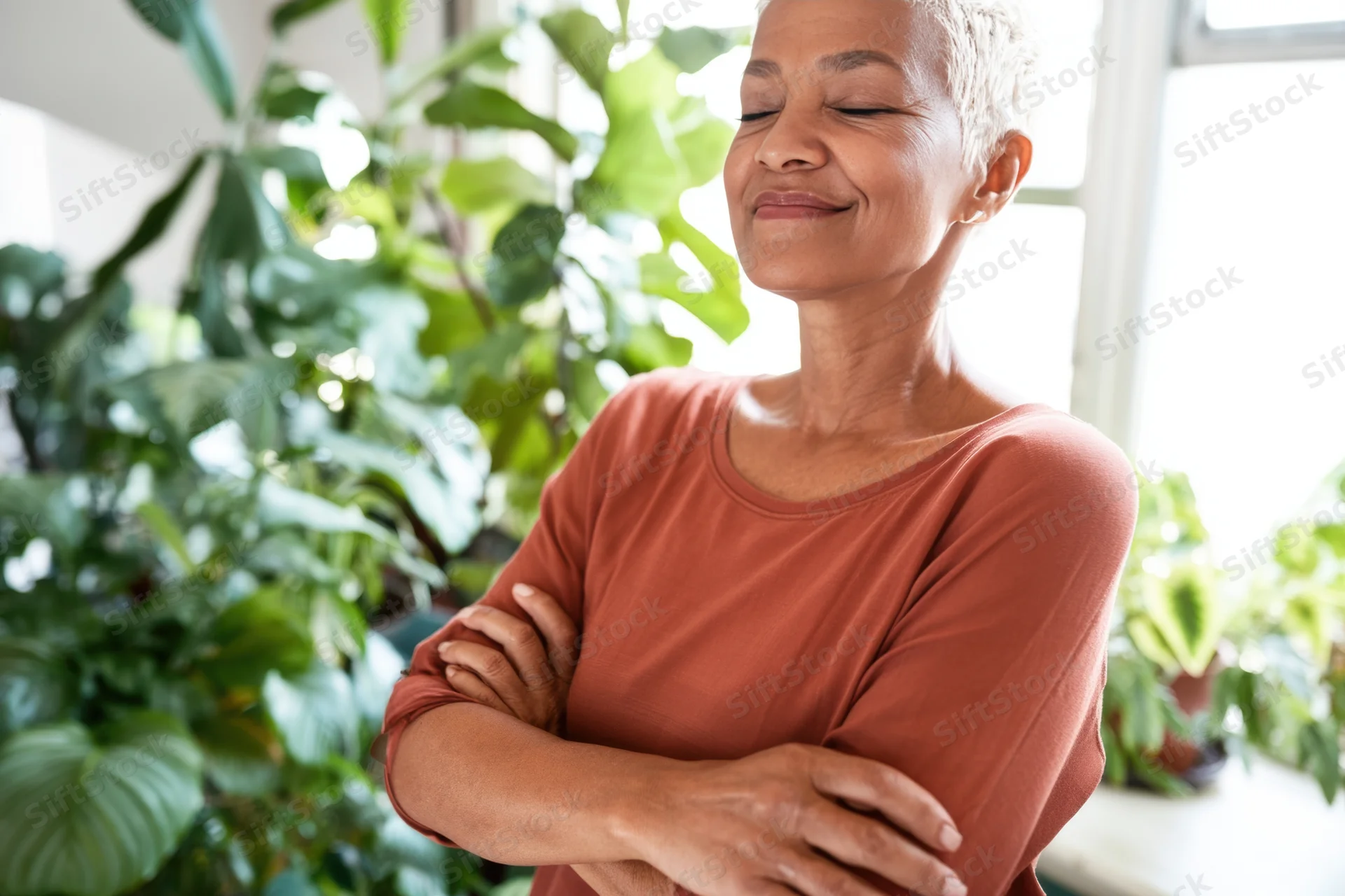 Serene woman with arms crossed smiling indoors near green plants
