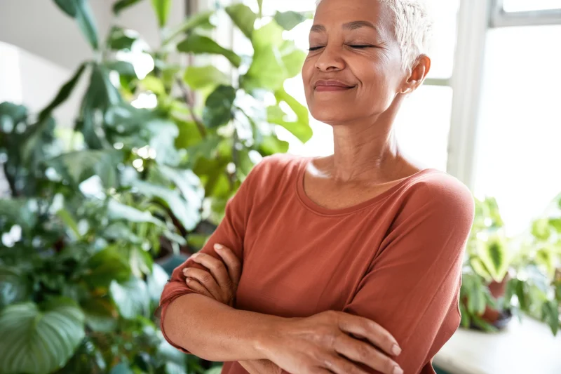 Serene woman with arms crossed smiling indoors near green plants