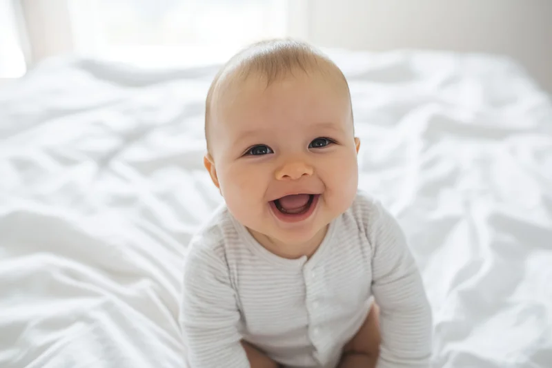 Smiling baby sitting up on a white bed wearing a striped onesie