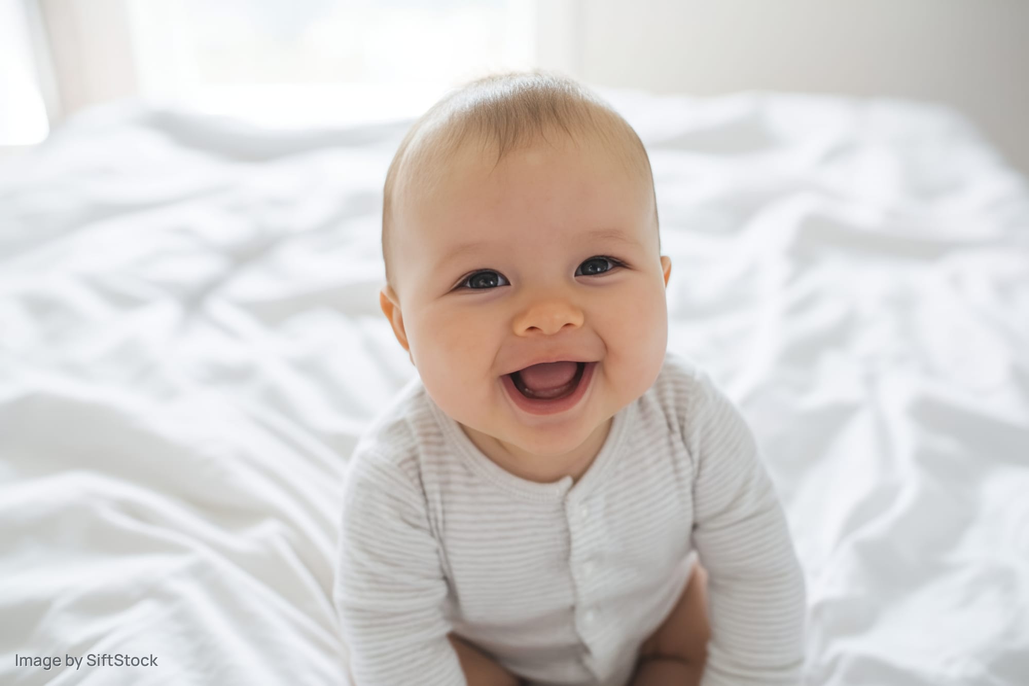 Smiling baby sitting up on a white bed wearing a striped onesie