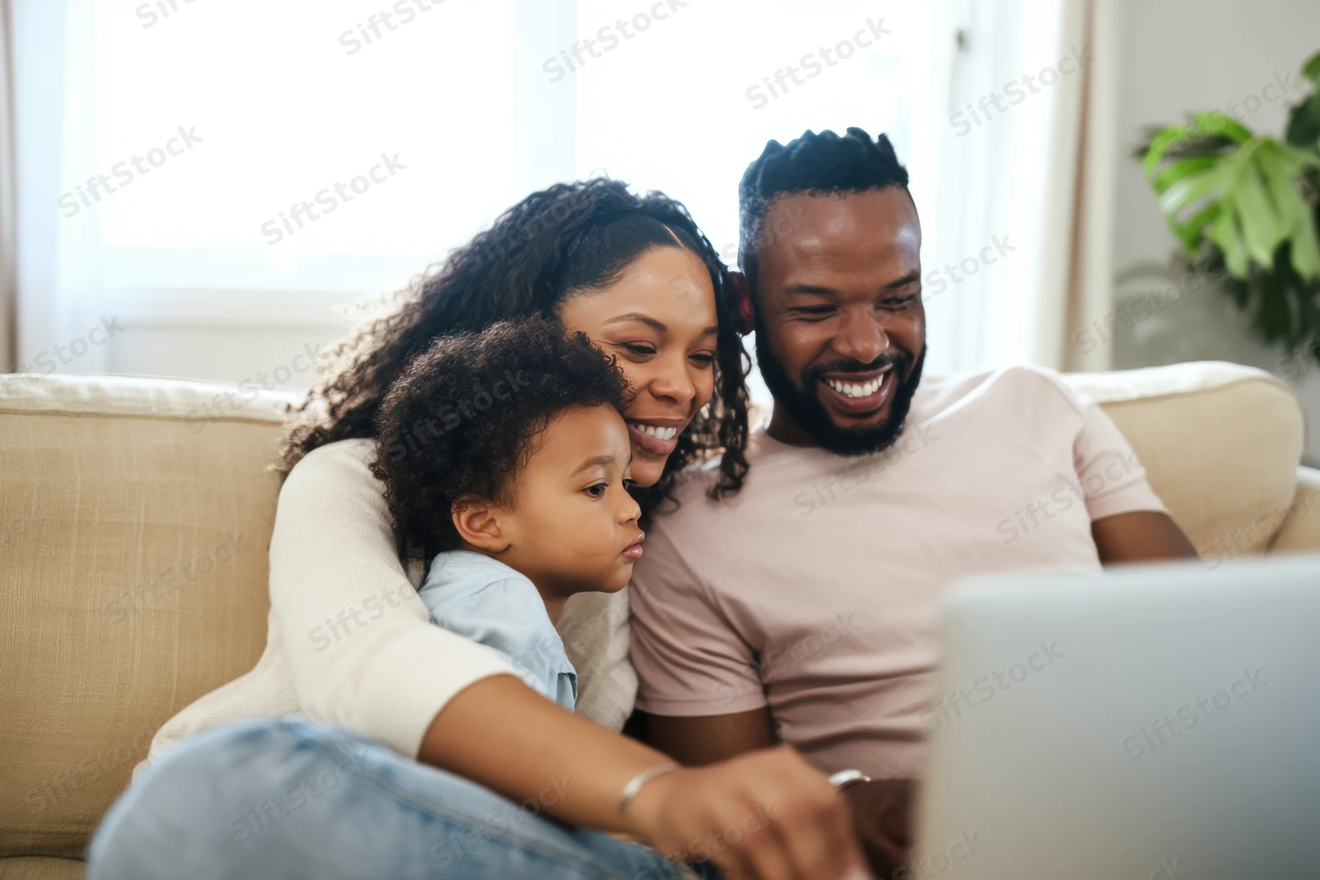 Black family smiling and laughing together on beige sofa while using laptop in bright living room