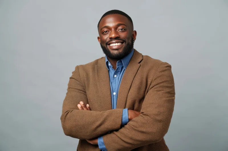 Smiling man with arms crossed in brown blazer over blue shirt against neutral background