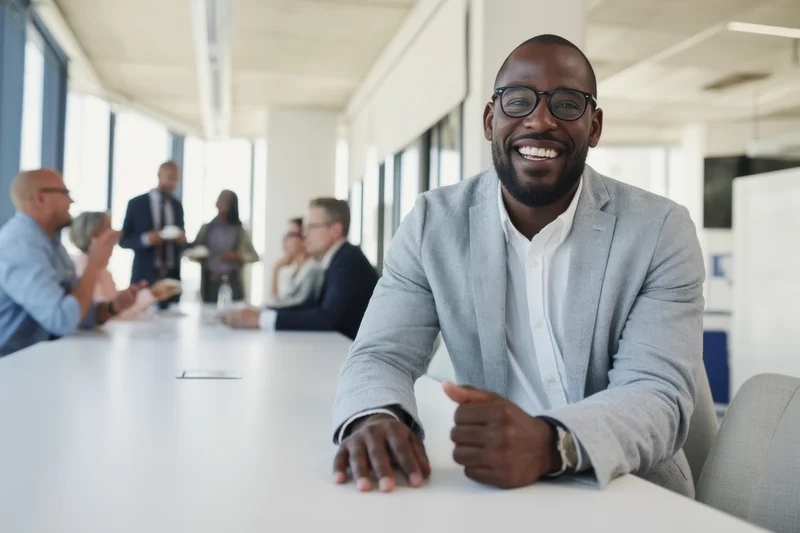 Smiling man in gray blazer sitting at conference table in modern office meeting with colleagues in background