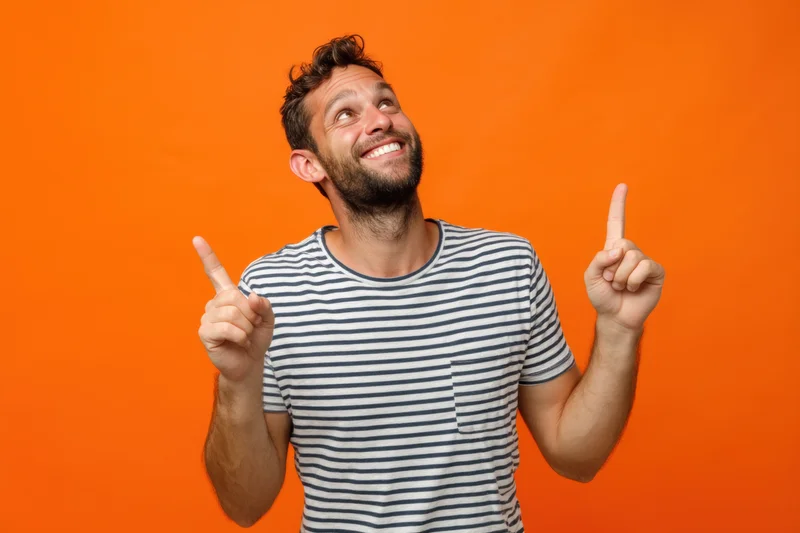 Smiling man in striped t-shirt pointing upward against bright orange background