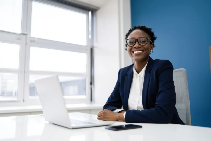 Smiling professional woman in navy blazer sitting at desk with laptop in office
