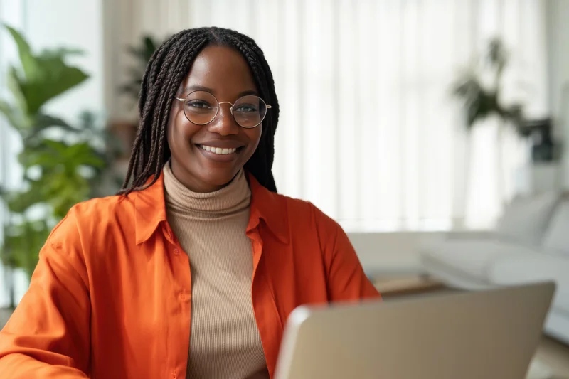 Smiling african woman with braided hair and round glasses wearing orange shirt and beige turtleneck using laptop at home