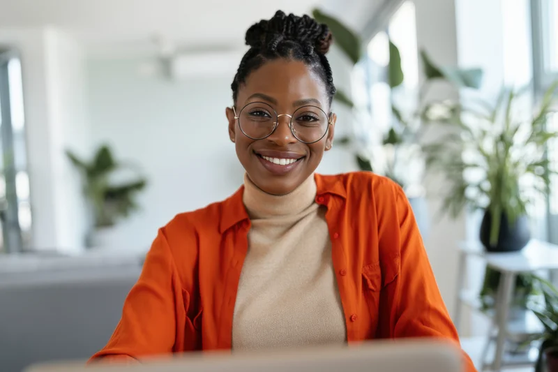 Smiling african woman with braided hair and round glasses sitting at laptop in bright modern office workspace
