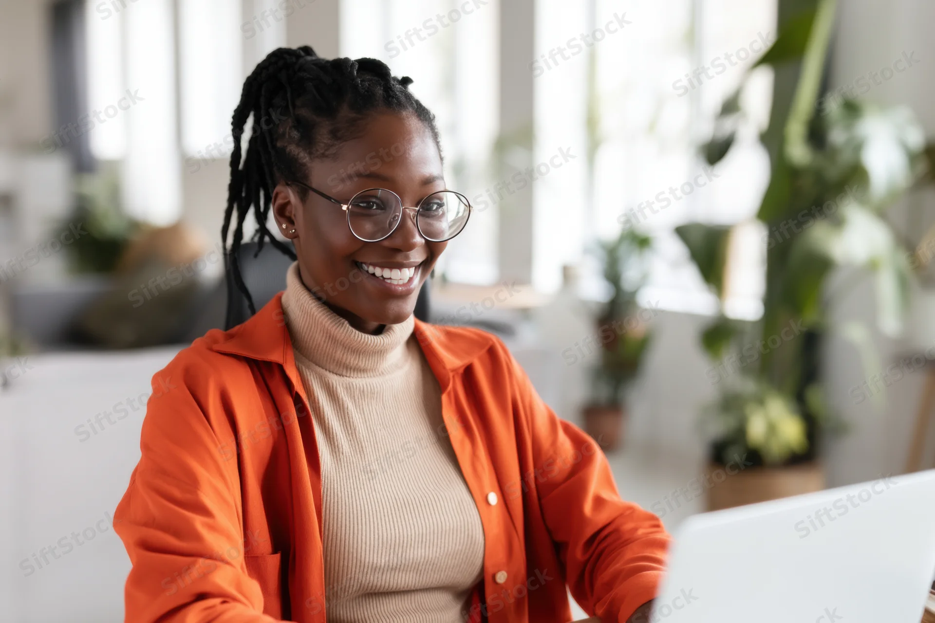 Smiling african woman in orange cardigan working on laptop in bright office with plants