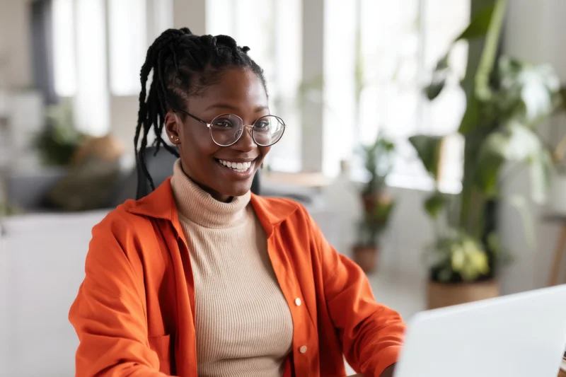 Smiling african woman in orange cardigan working on laptop in bright office with plants