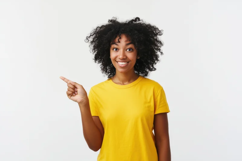 Smiling woman with curly black hair pointing left in yellow t-shirt against white background