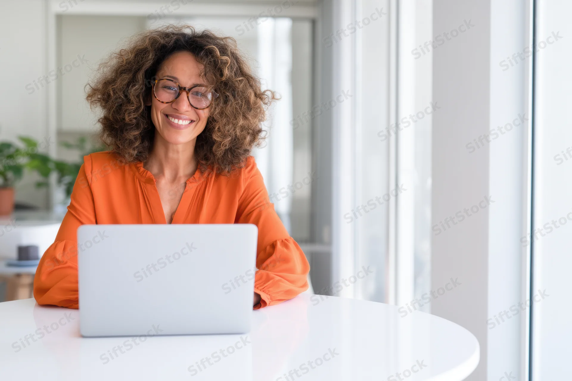 Smiling woman with curly hair and glasses wearing orange blouse using laptop at white desk in bright office space