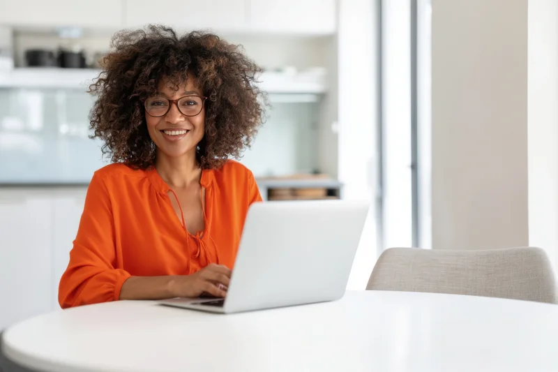 Smiling woman with curly hair and glasses working on laptop at kitchen table