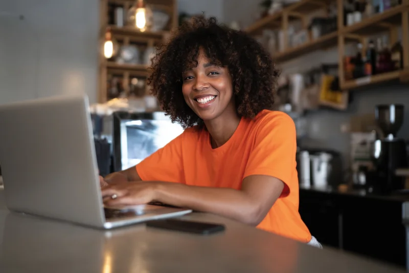 Smiling woman in orange t-shirt working on laptop at cafe counter
