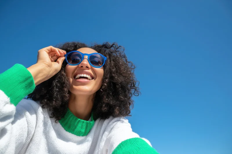 Smiling woman with curly hair wearing blue sunglasses and white green sweater outdoors under clear blue sky