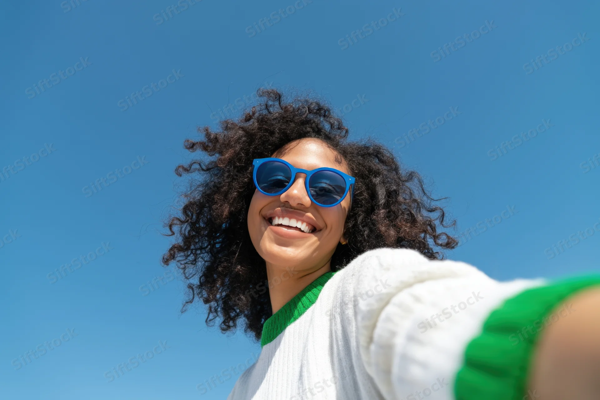Smiling woman with curly hair wearing blue sunglasses and white sweater outdoors under clear blue sky