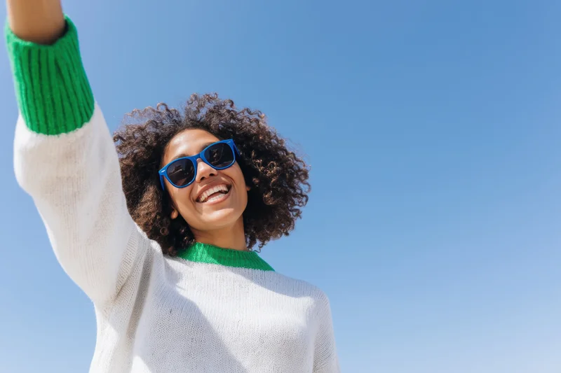 Smiling woman with curly hair wearing sunglasses and a white sweater with green trim, raising her arm against a clear blue sky