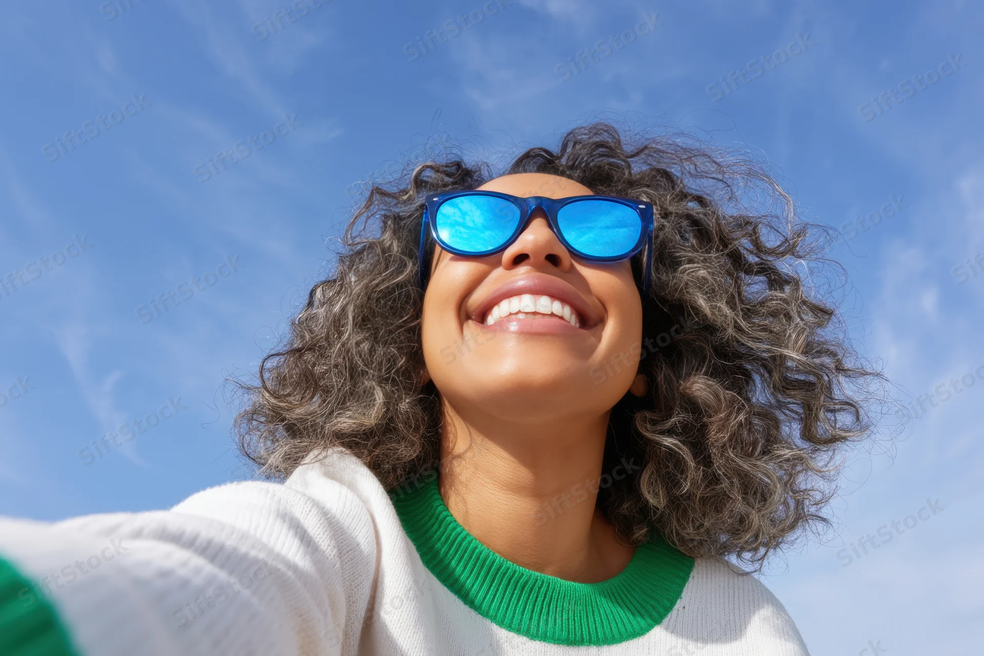 Smiling woman with curly hair wearing sunglasses and white sweater outdoors under blue sky