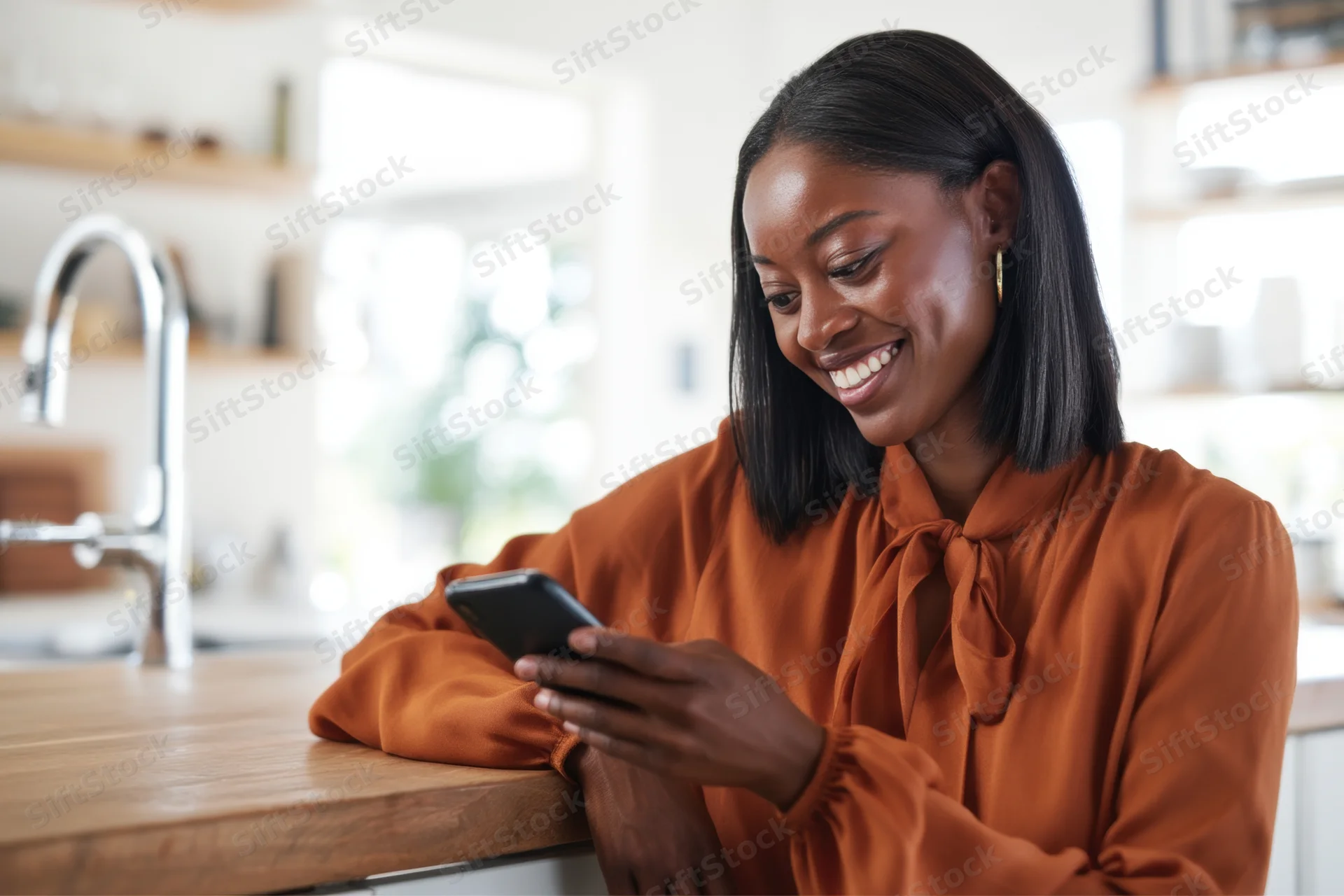 Woman smiling while using smartphone in kitchen leaning on wooden countertop