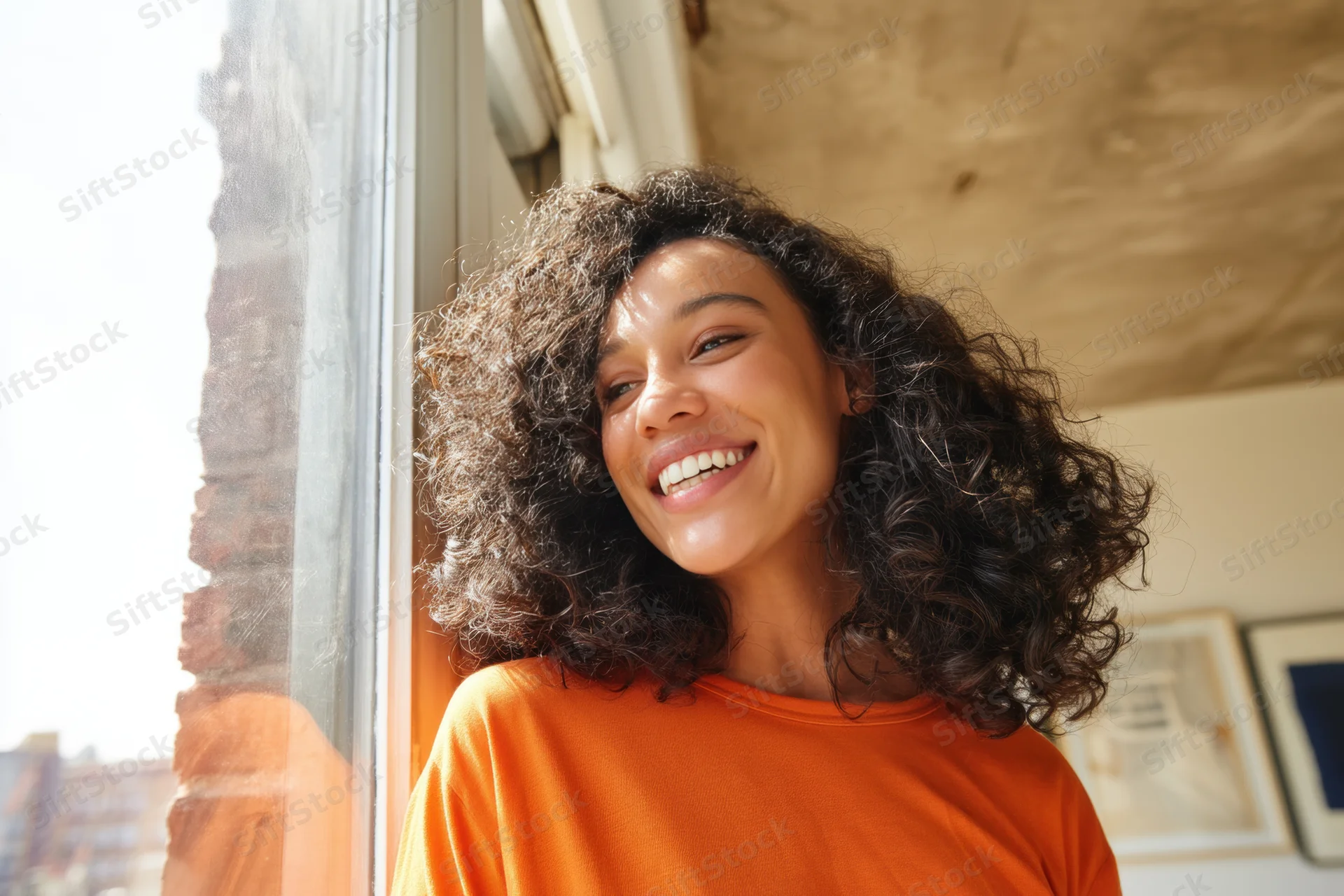 Young woman smiling with curly dark hair near window in orange shirt