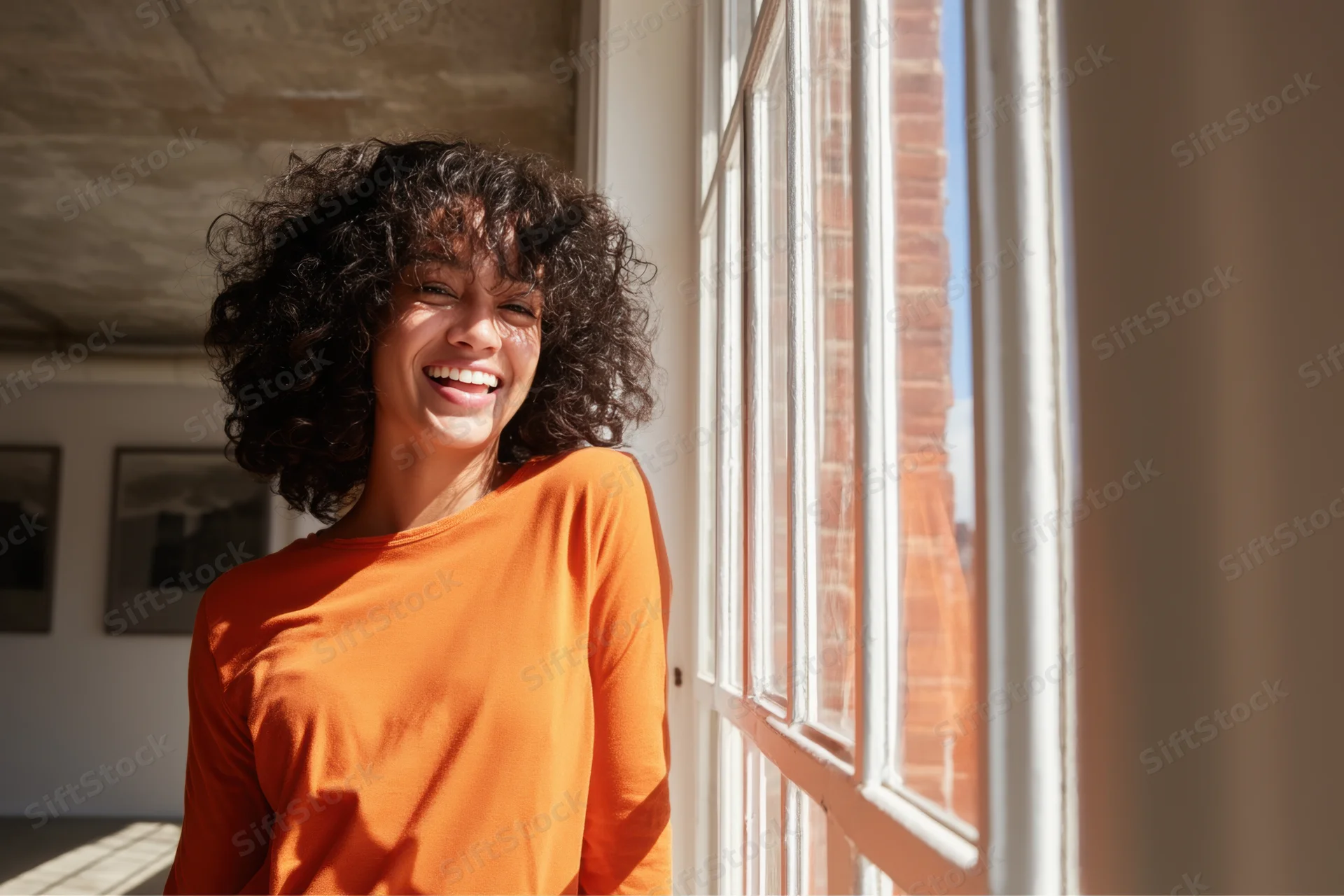 Young woman smiling by a window in a sunlit room wearing an orange shirt