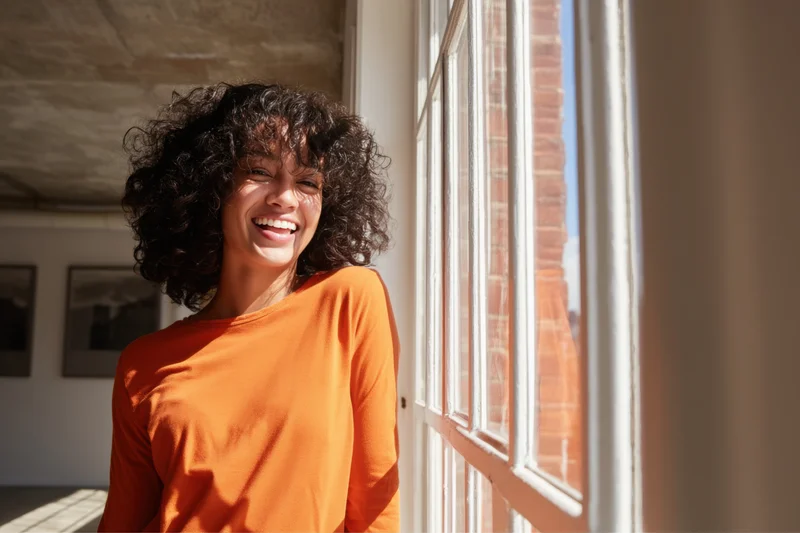 Young woman smiling by a window in a sunlit room wearing an orange shirt