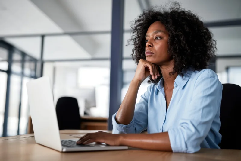 Woman sitting at desk with laptop, hand on chin, looking thoughtful in office setting