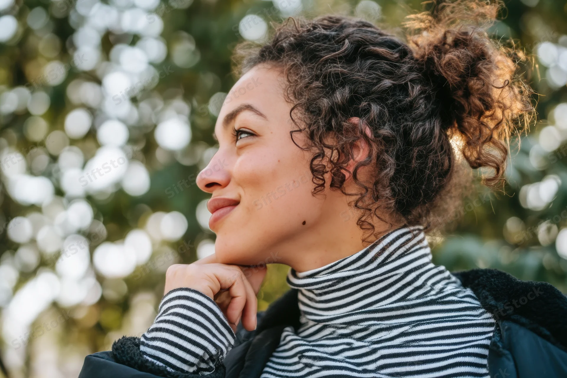 Woman with curly hair in a bun smiling thoughtfully outdoors in a striped turtleneck and jacket