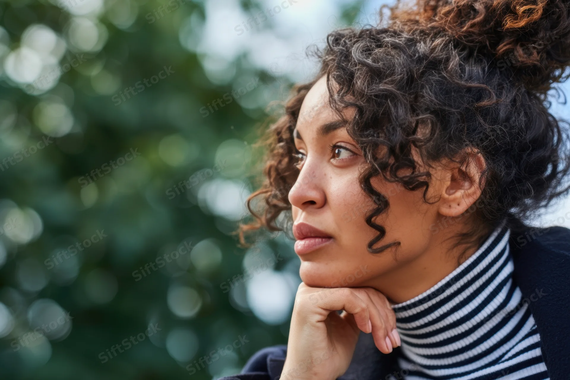 Thoughtful woman with curly hair in a bun resting her chin on her hand outdoors
