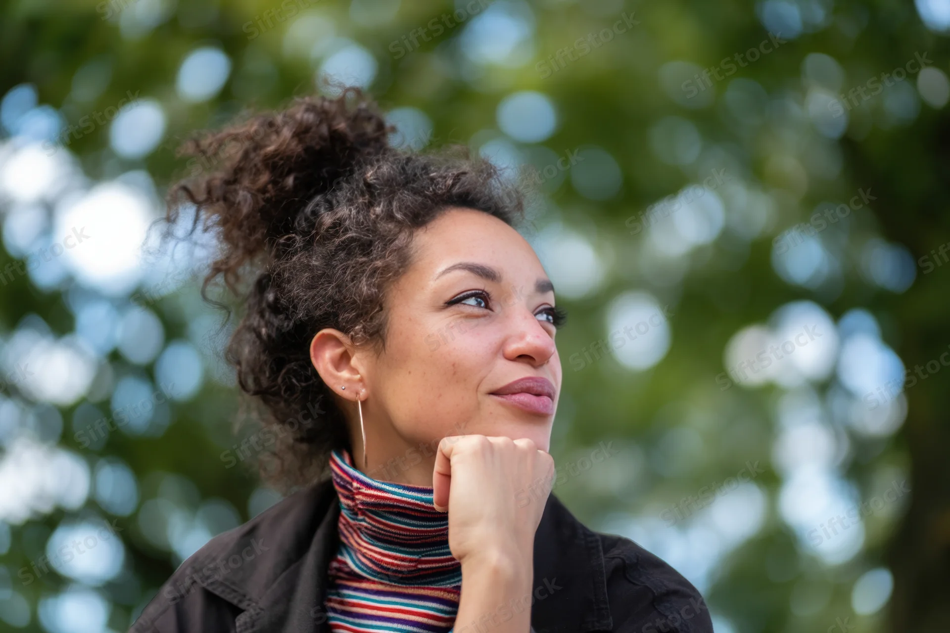 Thoughtful woman with curly hair in a bun, smiling gently while resting her chin on her hand outdoors