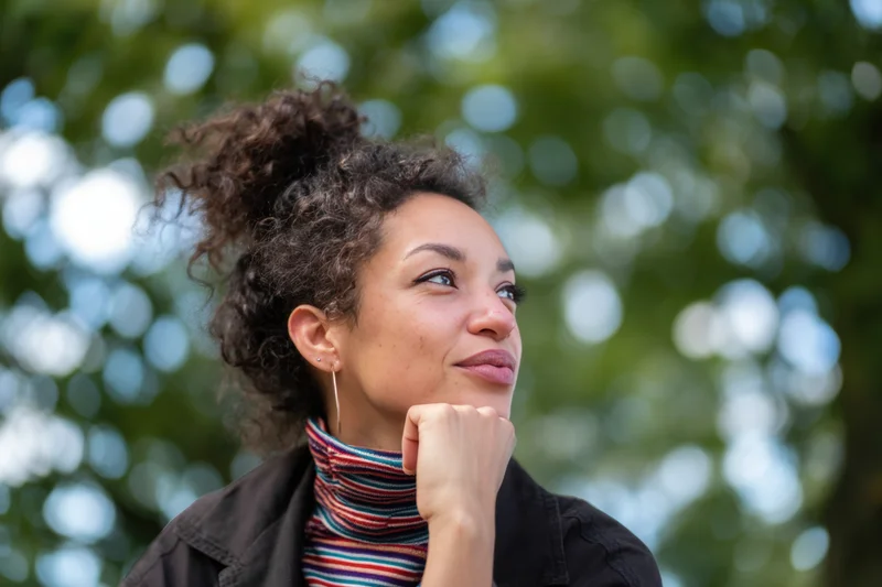 Thoughtful woman with curly hair in a bun, smiling gently while resting her chin on her hand outdoors
