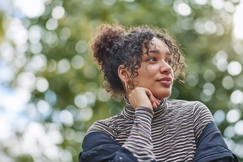 Thoughtful woman with curly hair in a bun, wearing a striped turtleneck, outdoors in a park with green foliage and sunlight