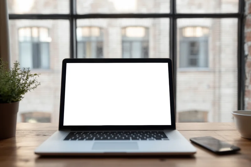 Silver laptop with blank white screen on wooden desk by window in modern office workspace