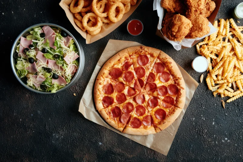 Top-down view of a fast food spread with pepperoni pizza, salad, onion rings, fries, and fried chicken on a dark textured surface