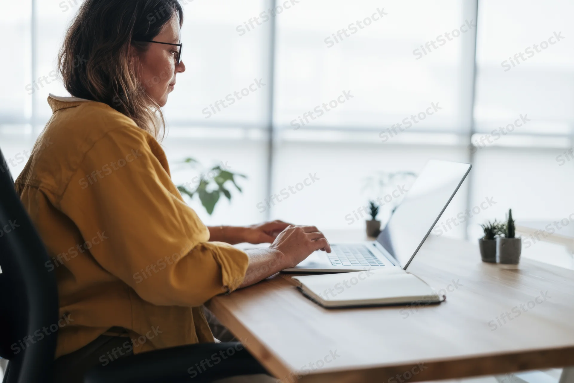 Woman working on laptop at wooden desk in bright office with plants and window view