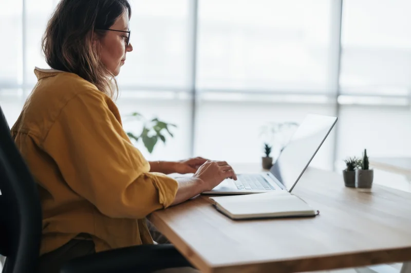 Woman working on laptop at wooden desk in bright office with plants and window view